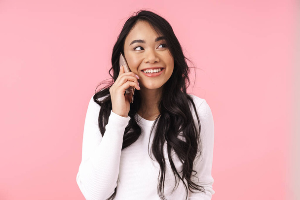 Image of young brunette asian woman with long hair talking on cellphone isolated over pink background - Photo, Image