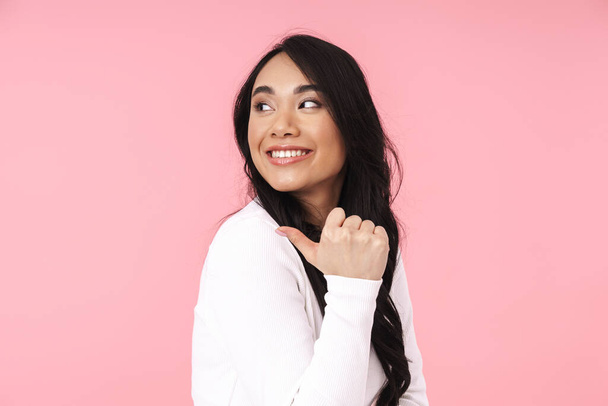 Image of young brunette asian woman in casual clothes smiling and pointing fingers aside isolated over pink background - 写真・画像