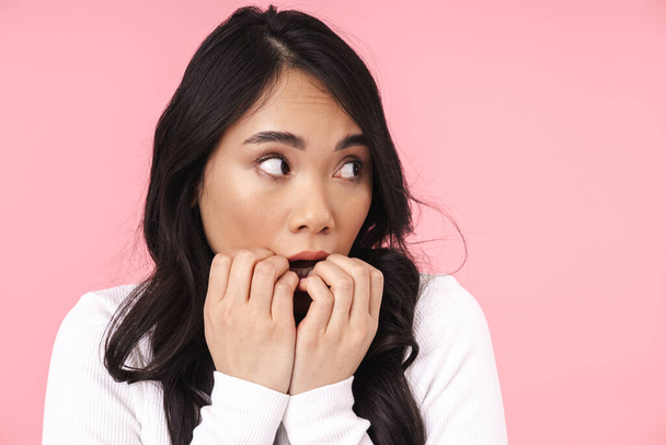 Image of young brunette asian woman in casual clothes being scared and biting her nails isolated over pink background - Photo, Image