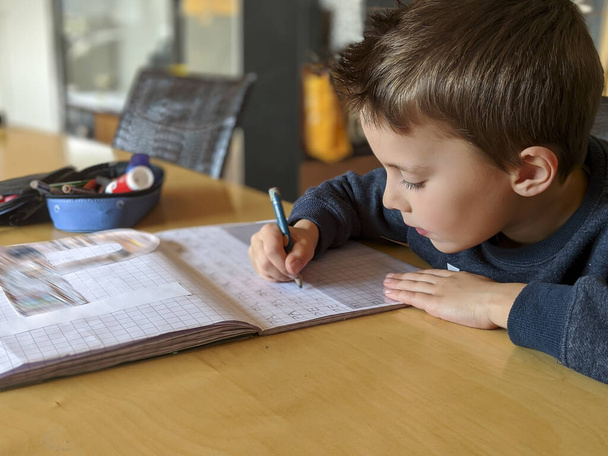 Portret van ijverig klein kind dat zorgvuldig aan het bureau zit en huiswerk maakt - Foto, afbeelding