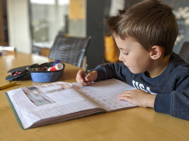Portrét pilného malého dítěte, jak pečlivě píše za stolem a dělá domácí úkoly. - Fotografie, Obrázek