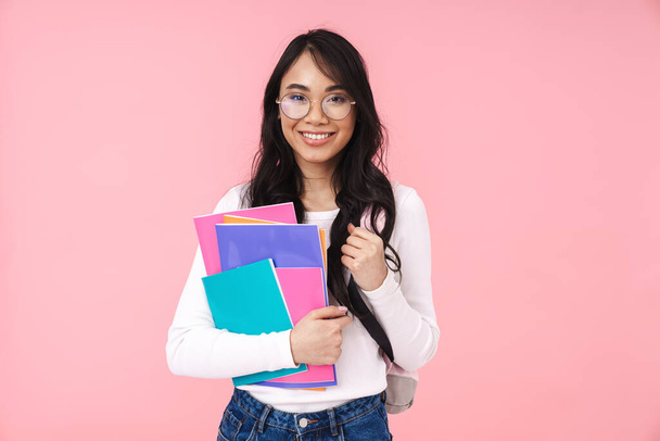 Image of young brunette asian student girl wearing eyeglasses holding paper folders isolated over pink background - Foto, Imagem