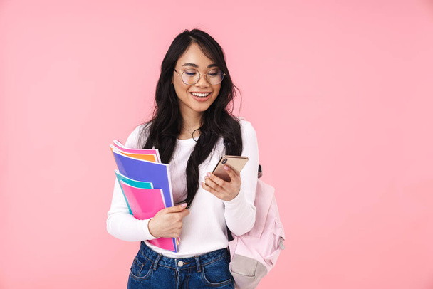 Image of young brunette asian student girl wearing eyeglasses holding paper folders isolated over pink background - Fotó, kép