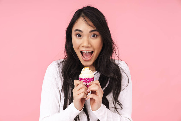 Image of young brunette asian woman with long hair smiling and holding cupcake isolated over pink background - Foto, afbeelding