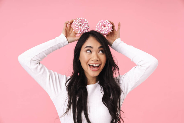 Image of young brunette asian woman with long hair smiling and holding donuts isolated over pink background - Foto, afbeelding