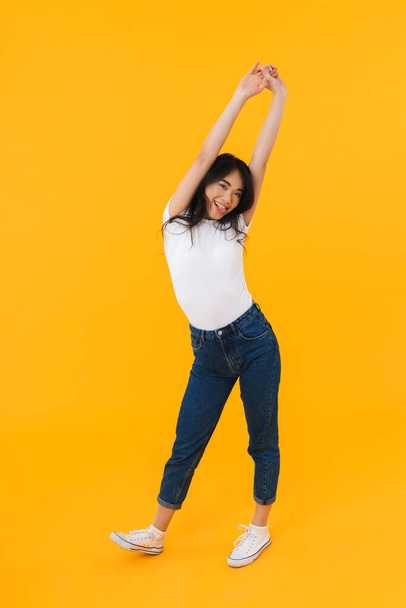 Full length image of young brunette asian woman wearing casual clothes smiling and having fun isolated over yellow background - 写真・画像