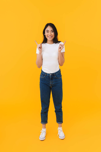 Full length image of young brunette asian woman smiling and pointing fingers at copyspace isolated over yellow background - Fotografie, Obrázek