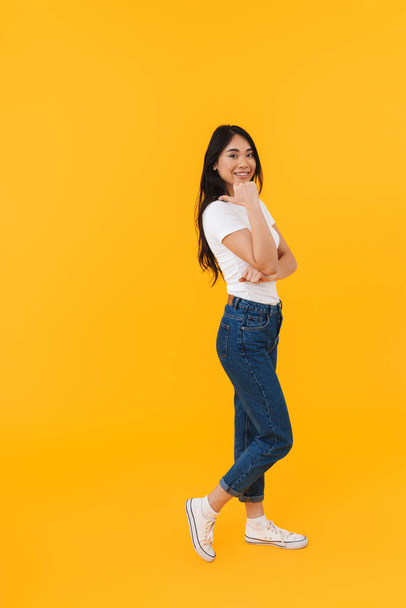 Full length image of young brunette asian woman smiling and pointing finger at copyspace isolated over yellow background - Фото, зображення