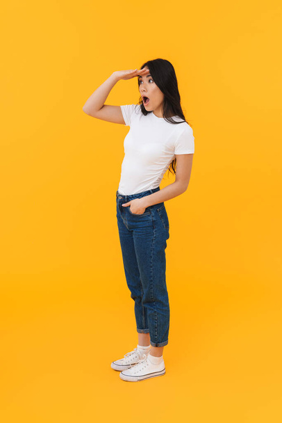Full length image of young brunette asian woman wondering and looking in distance at copyspace isolated over yellow background - 写真・画像