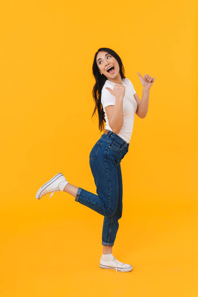 Full length image of young brunette asian woman smiling and pointing fingers at copyspace isolated over yellow background - Fotó, kép