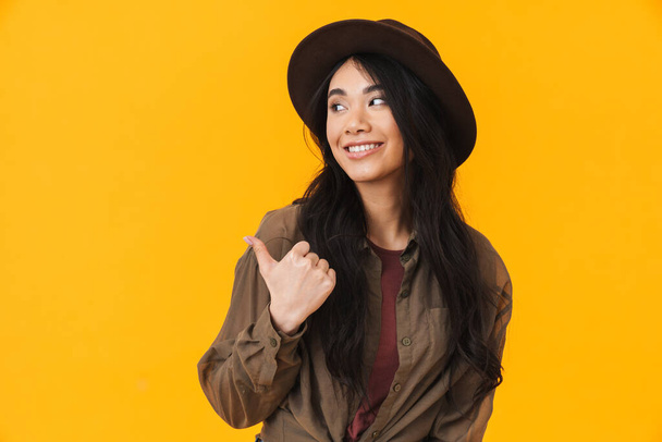 Image of young brunette asian woman wearing hat smiling and pointing finger at copyspace isolated over yellow background - Foto, afbeelding
