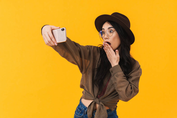 Image of young brunette asian woman wearing hat and eyeglasses taking selfie photo on cellphone isolated over yellow background - Zdjęcie, obraz