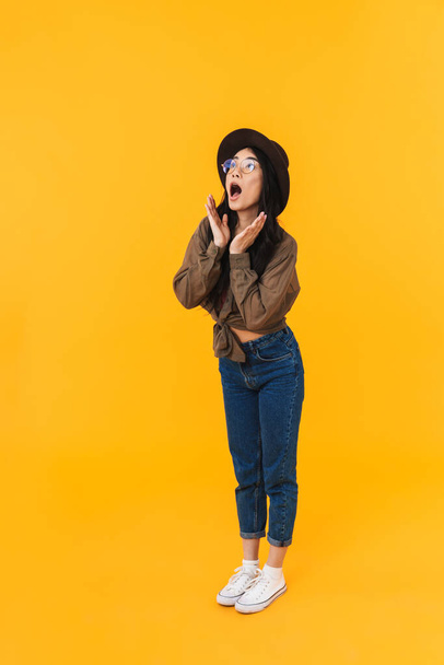 Full length image of young brunette asian woman wearing hat expressing surprise and looking at copyspace isolated over yellow background - Фото, зображення