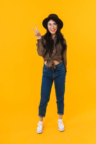 Full length image of young brunette asian woman wearing hat smiling and gesturing peace fingers isolated over yellow background - Фото, зображення