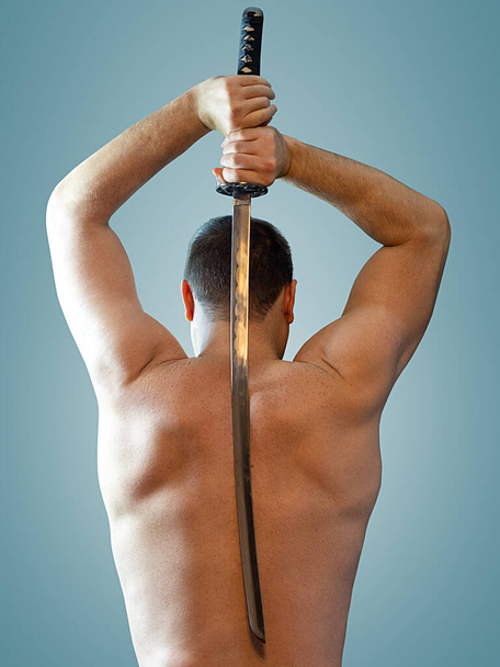 View from the back of a muscular man with a naked torso holding a sword behind his back on an isolated background - Photo, Image