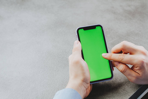 Man touches and looks at the screen of the smartphone with a green screen. Chroma key on the screen of the telephone in businessman's hands on gray background - Photo, Image