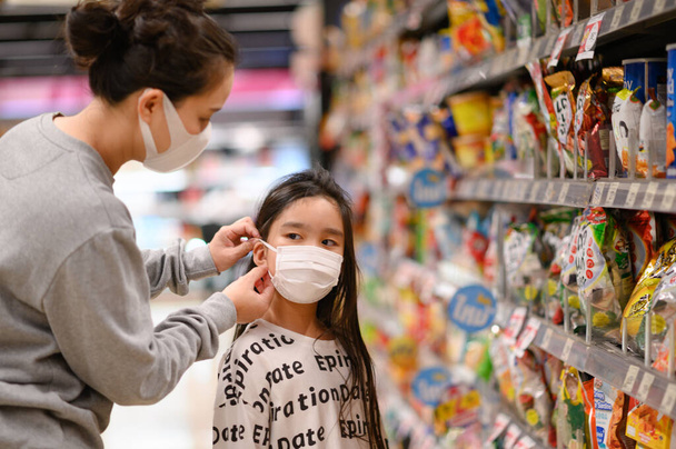 Una mujer que lleva una máscara protectora le pone una máscara facial a una hija en el supermercado. Seguridad durante el brote de COVID-19. Epidemia de virus covid, tomada en Ramintra, Tailandia, en marzo de 2020
. - Foto, Imagen