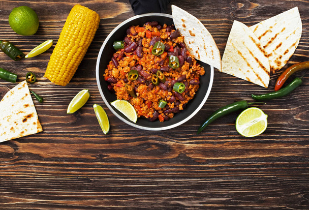 Παραδοσιακό μεξικάνικο τσίλι con carne σερβίρεται σε ρουστίκ ξύλινο φόντο σε ένα τηγάνι με καλαμπόκι, μεξικάνικη τορτίγια ψωμί, ασβέστη και jalapeno. Στο πάνω μέρος. Χώρος για κείμενο - Φωτογραφία, εικόνα