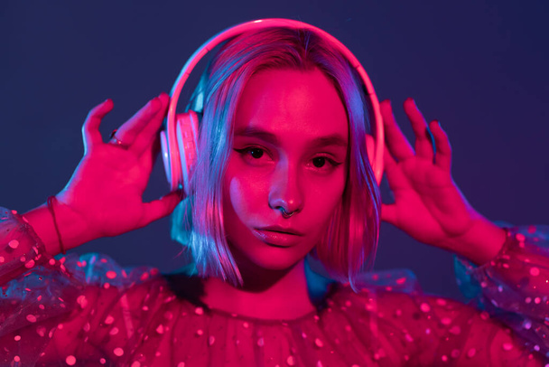 Gorgeous lady with dyed hair listening music in headphones and singing with neon light background. Charming hipster girl dancing with eyes closed. - Foto, Imagen