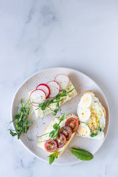 Breakfast sandwiches with cream cheese, quail eggs, tomatoes, radish and micro green on a ceramic plate, light grey background. Healthy snack. Top view.  - Foto, Imagen