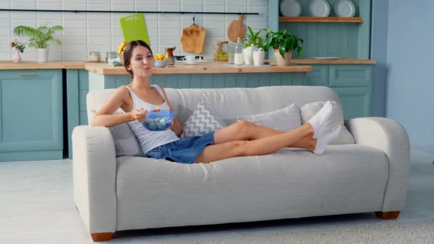 Cute girl eating fruit salad sitting on sofa - Filmati, video