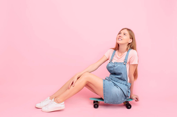 Happy attractive stylish young woman sitting on a skateboard on an isolated pink background - Фото, зображення