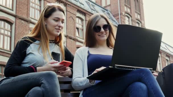Moderne Studentinnen arbeiten gemeinsam an einem Laptop an einem Studentenprojekt, während sie vor dem Unterricht auf einer Bank im Campus-Park sitzen. Hochschulbildung - Filmmaterial, Video