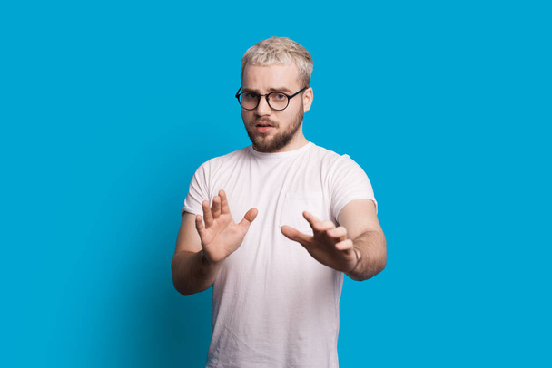 Blanke man met bril en blond haar is gebaren angst op een blauwe achtergrond - Foto, afbeelding