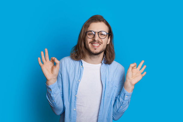 Homem caucasiano de cabelos longos com barba e óculos está gesticulando o sinal ok em uma parede azul
 - Foto, Imagem