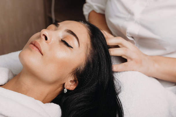 Close up portrait of a caucasian woman with black hair lying on couch during a head massage procedure - Photo, Image
