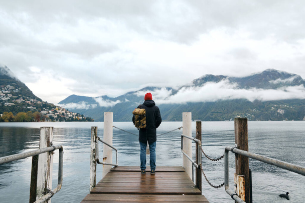 Man with backpack enjoy panorama on pier in Lugano. Man in travel. Lake Lugano, southern slope of Alps. Landscape in Switzerland. Amazing scenic outdoors view. Canton of Ticino. Adventure lifestyle - Φωτογραφία, εικόνα