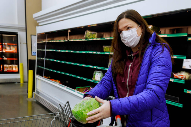 Mujer con máscara facial comprando lechuga en el supermercado con estantes vacíos. Concepto pandémico del Coronavirus
. - Foto, Imagen