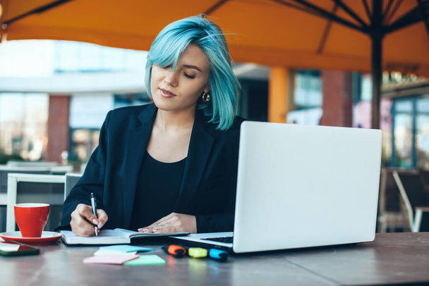 Καυκάσιος επιχειρηματίας με μπλε μαλλιά κάνει κάποιες σημειώσεις σε μια καφετέρια χρησιμοποιώντας έναν υπολογιστή - Φωτογραφία, εικόνα