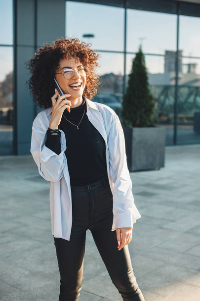 Curly haired business person talking on phone while smiling and wearing glasses outside - Zdjęcie, obraz