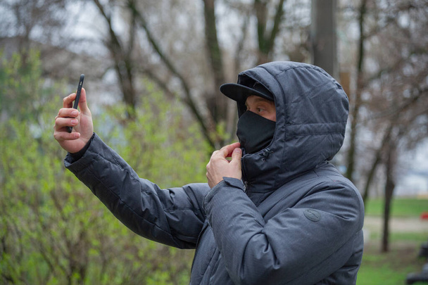 Egy férfi fekete légzőmaszkot visel az arcán. Koronavírus. Egy fickó tavasszal az utcán, légzőmaszkban, hogy védekezzen a vírusfertőzés ellen. Egy férfi lefényképezi magát egy okostelefonon.. - Fotó, kép