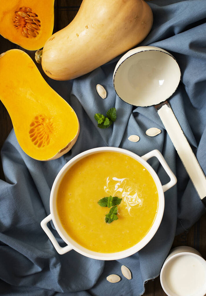 Autumn Pumpkin Cream soup on a wooden background with a gray tablecloth, served with cream, pumpkin seeds and mint. Cut pumpkin, soup ladle, white plate. Top view. Vertical - Photo, Image