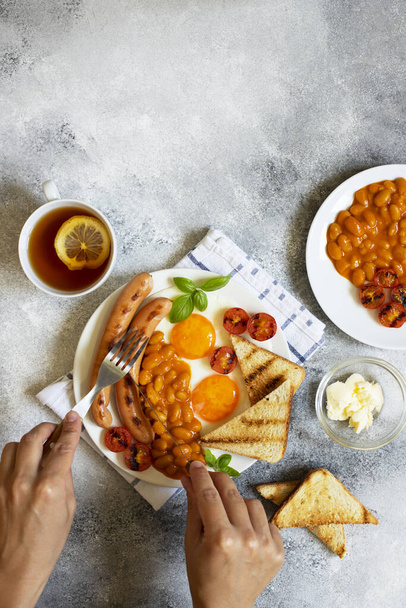 Female hands as she is having English breakfast with fried sausages, beans, mushrooms, fried eggs, grilled tomatoes. Served with a cup of tea with lemon, bread toast and butter. Gray background - Foto, immagini