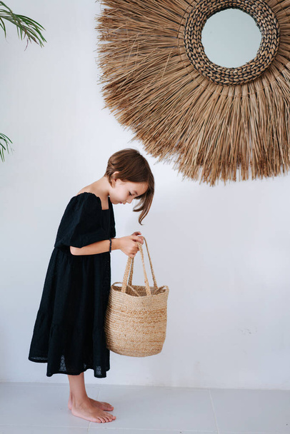 Cute barefoot little girl in a midi black dress standing with a wicker handbag in a tropical style room. She's looking inside it. Side view. - Foto, afbeelding