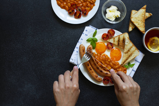 Mains féminines comme elle prend son petit déjeuner anglais avec des saucisses frites, haricots, champignons, œufs frits, tomates grillées. Servi avec une tasse de thé au citron, pain grillé et beurre. Fond noir - Photo, image
