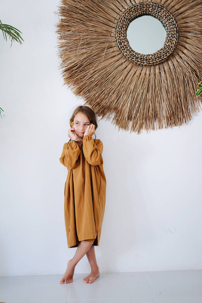 Smiling barefoot little girl in an ochre midi dress worn over white shirt posing for a photo in a tropical style room. She's holding hair tassels in hands, looking to side. - Fotografie, Obrázek