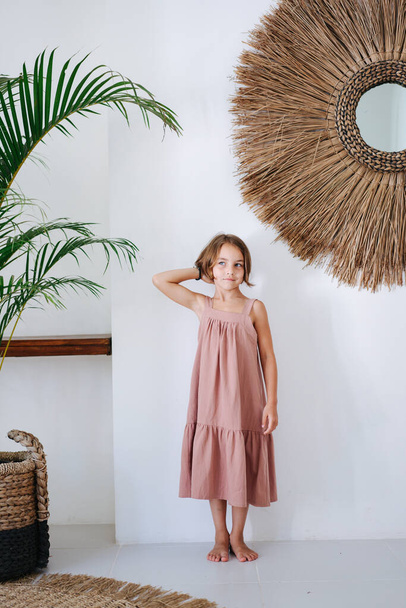 Cute barefoot little girl in a pink midi dress posing for a photo in a tropical style room. Her hand on a back of her head. - Photo, Image