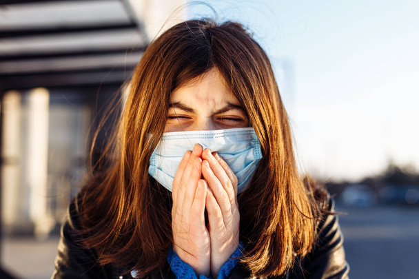 Mladá dívka kýchne na autobusovém nádraží a zakryje si obličej rukama. Dívka nosí bílou lékařskou masku, která stojí poblíž autobusu na zastávce veřejné dopravy. Koncept koronavirové nemoci. Pandemie, epidemie - Fotografie, Obrázek