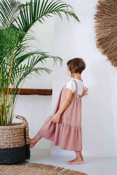 Cute barefoot little girl in a pink midi dress worn over white in a tropical style room. She's pushing heavy potted plant with her foot. Side view. - Foto, afbeelding