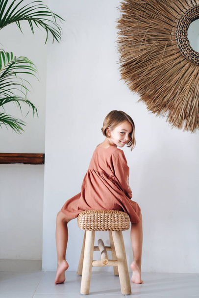 Roztomilá bosá holčička v tmavě oranžových šatech, pózuje pro fotografii v místnosti v tropickém stylu. Sedí na proutěné lavičce, nohy na každé straně. Zpětný pohled na kameru. - Fotografie, Obrázek