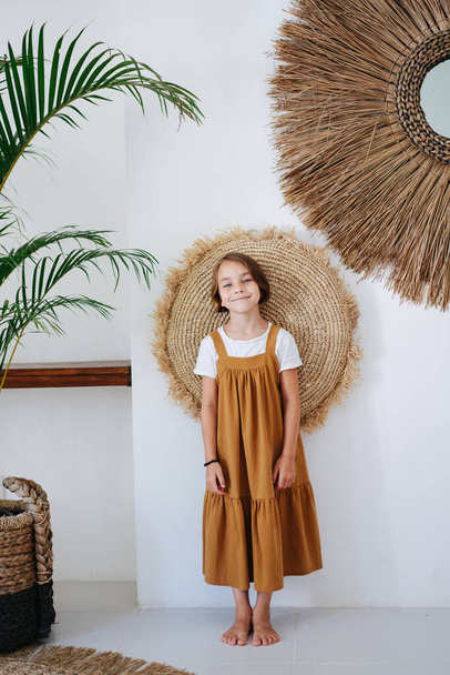 Smiling barefoot little girl in an ochre midi dress worn over white shirt posing for a photo in a tropical style room. She's leaning her back on a pillow, pressing it to the wall. - Foto, immagini