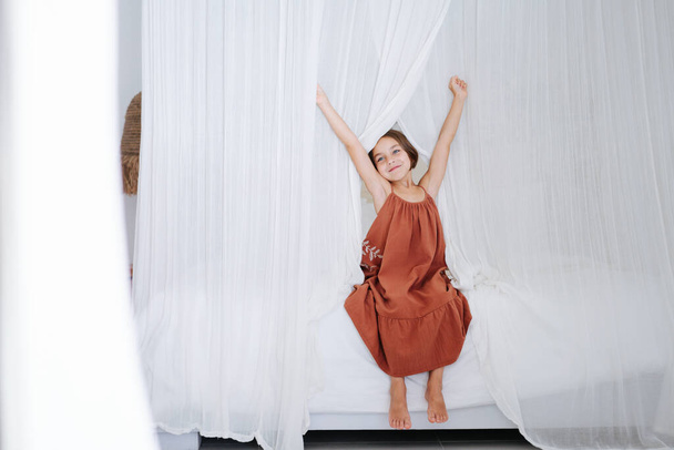 Cute barefoot little girl in a short dark orange dress, sitting, stretching on a double bed with white curtains. Looking to side. - Foto, immagini