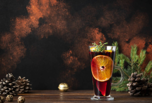 Glühwein, ein warmes Getränk aus Rotwein, Zitrusfrüchten und Gewürzen auf einem Holztisch mit Weihnachtsdekoration. Glas und Karaffe mit Glühwein. Dunkler Hintergrund. Nahaufnahme, bunt, Kopierraum - Foto, Bild