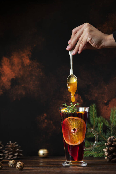Mulled wine a warm drink made of red wine, citruses and spices in a glass on a wooden table with Christmas decorations. Female hand putting honey from a spoon into mulled wine. Dark background - Photo, Image