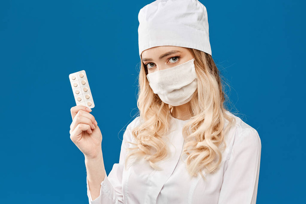 Young woman doctor in white coat and medical mask showing pack of pills, isolated at a blue background with copy space. Nurse in white medical uniform and protective mask keeps pills in her hand - Zdjęcie, obraz
