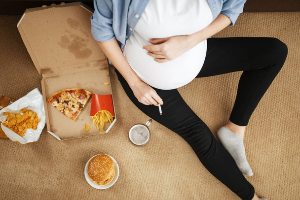 Pregnant woman with belly smoking and eats unhealthy food at home. Pregnancy, bad habits and lifestyle in prenatal period. Ugly expectant mom, health damage - Foto, afbeelding
